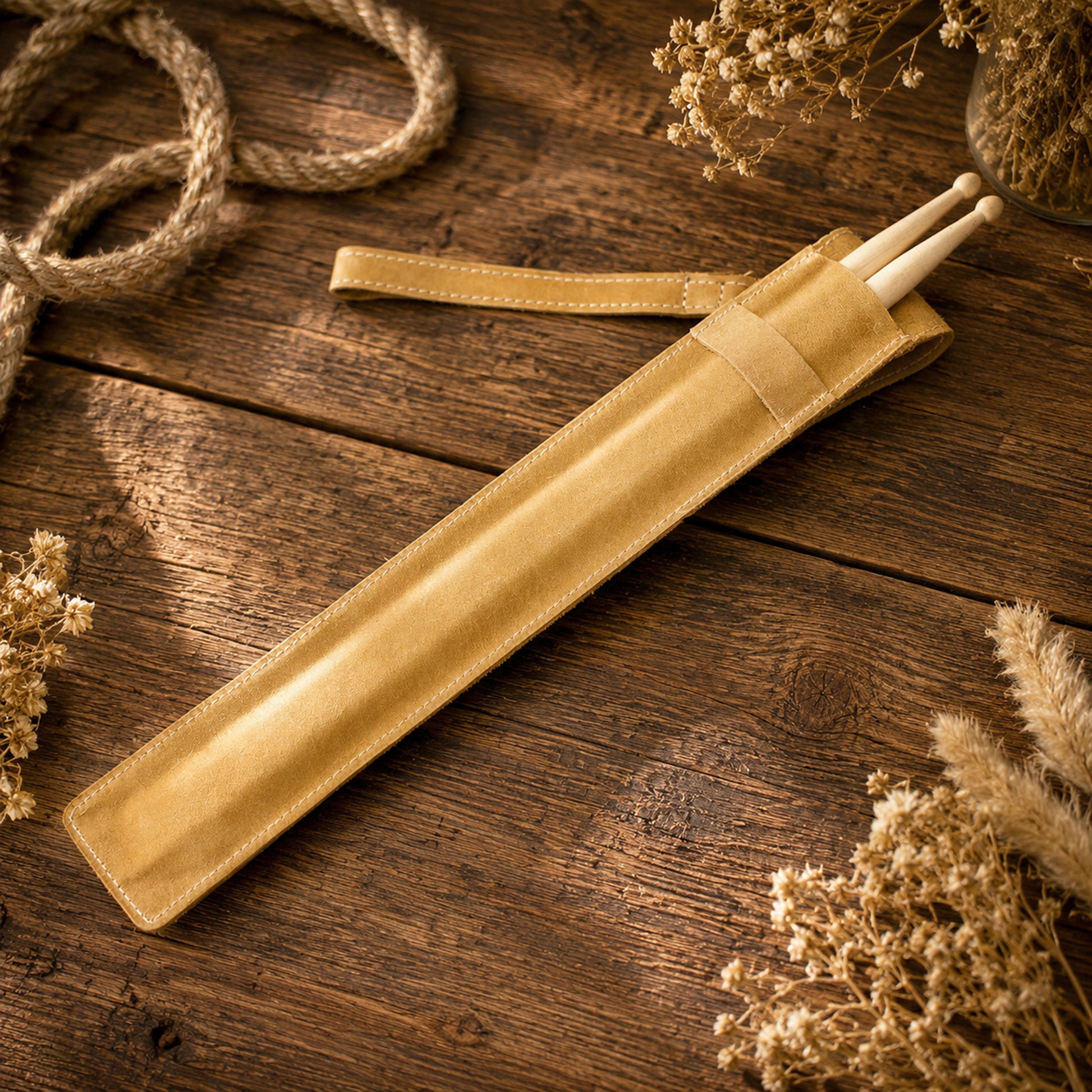 A tan leather drumstick sleeve holding two light wood drumsticks, arranged diagonally on a rustic dark wood tabletop with dried flowers and rope as styled props.