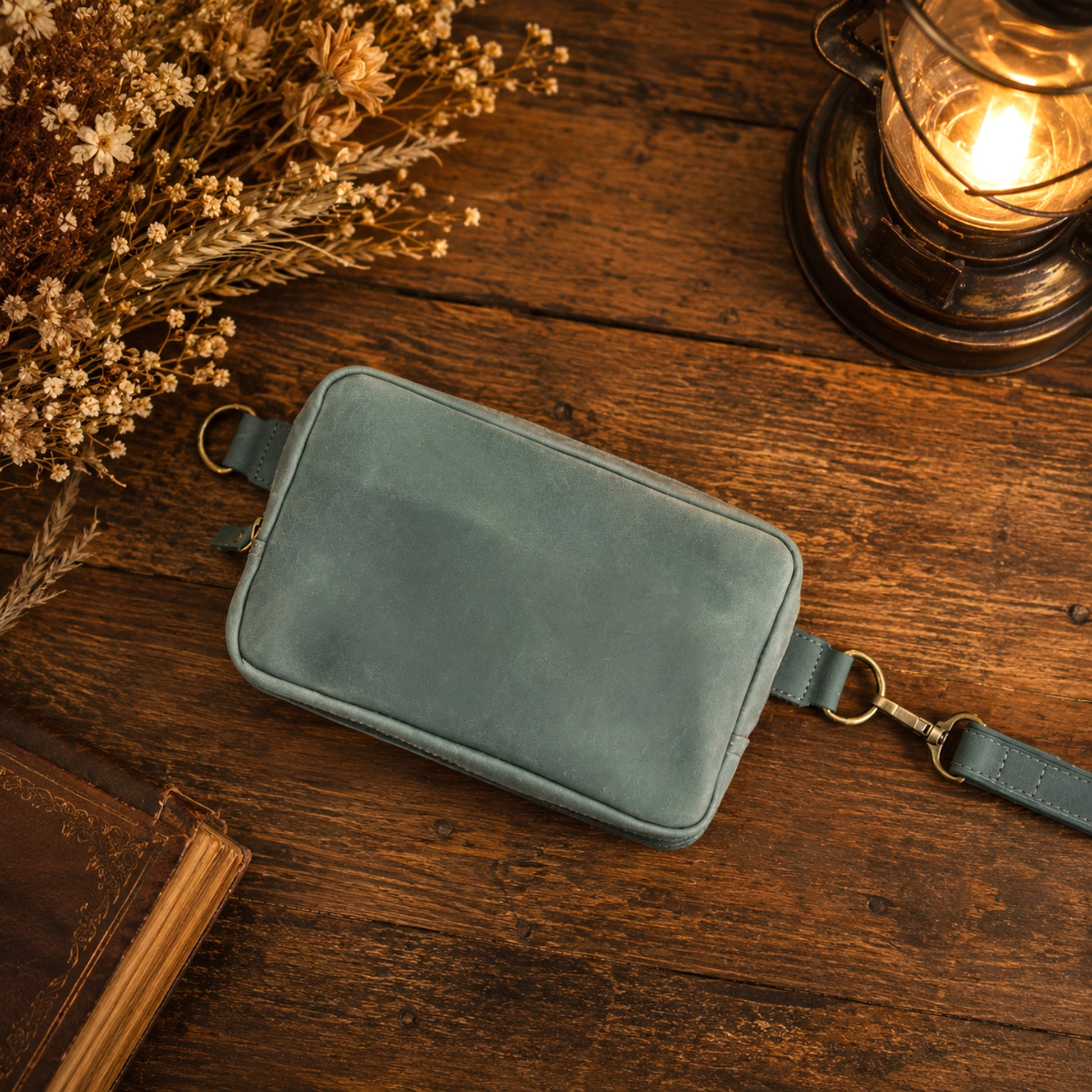 A dusty blue-green leather crossbody pouch with gold-tone hardware, styled on a rustic wooden table next to dried flowers, a book, and a glowing vintage lantern.