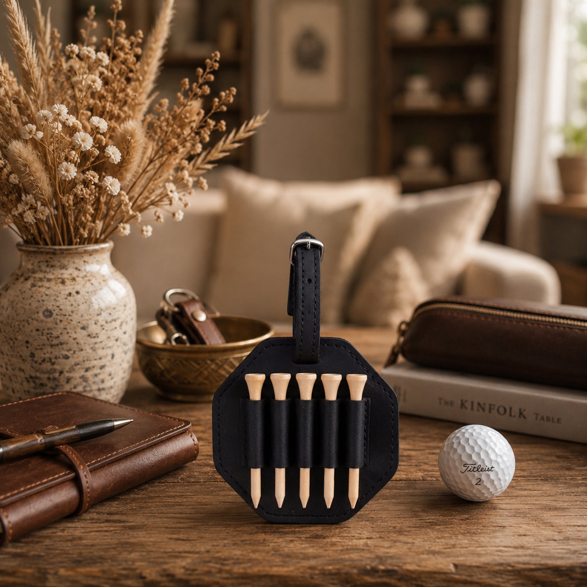 A black leather golf teiger set holder with five wooden teiger sticks, displayed on a rustic wooden table next to a golf ball, a brown leather notebook, and a vase of dried grasses in a cozy living room setting.