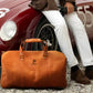Orange duffel bag with a brand logo in front of a red vintage car.