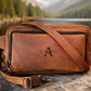 Brown leather bag with a visible brand logo on a rock by a lake with mountains in the background