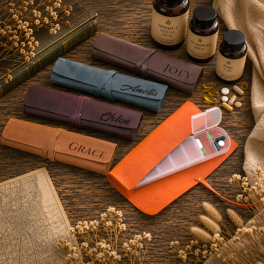 A styled flat-lay of five personalized pill organizer rolls in different colors (brown, teal, burgundy, tan, and orange) with names engraved on them, alongside supplement bottles and a clear plastic pill organizer tray with open compartments, all arranged on a rustic wooden table with dried flowers and a vintage map.