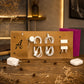 A tan leather organizer stand with multiple elastic loops and slots holding white charging cables, adapters, and earbuds, displayed on a wooden table with dried flowers and blurred background decor.