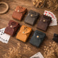 Six small leather card cases arranged on a rustic wooden surface with playing cards, dried flowers, rope, and a glass jar in the background. The cases are in brown, gray, burgundy, mustard yellow, and dark teal, each with an embossed monogram or name and a snap closure.