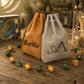 Two personalized drawstring pouches on a rustic wooden table, surrounded by colorful polyhedral gaming dice. The left pouch is tan with the name 'Bertie' printed in black, and the right pouch is light gray with a black monogram logo.
