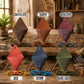 Six diamond-shaped leather accessory holders in different colors are arranged on a rustic wooden table. Each holder has a metal stand, a strap, and a small round snap closure.