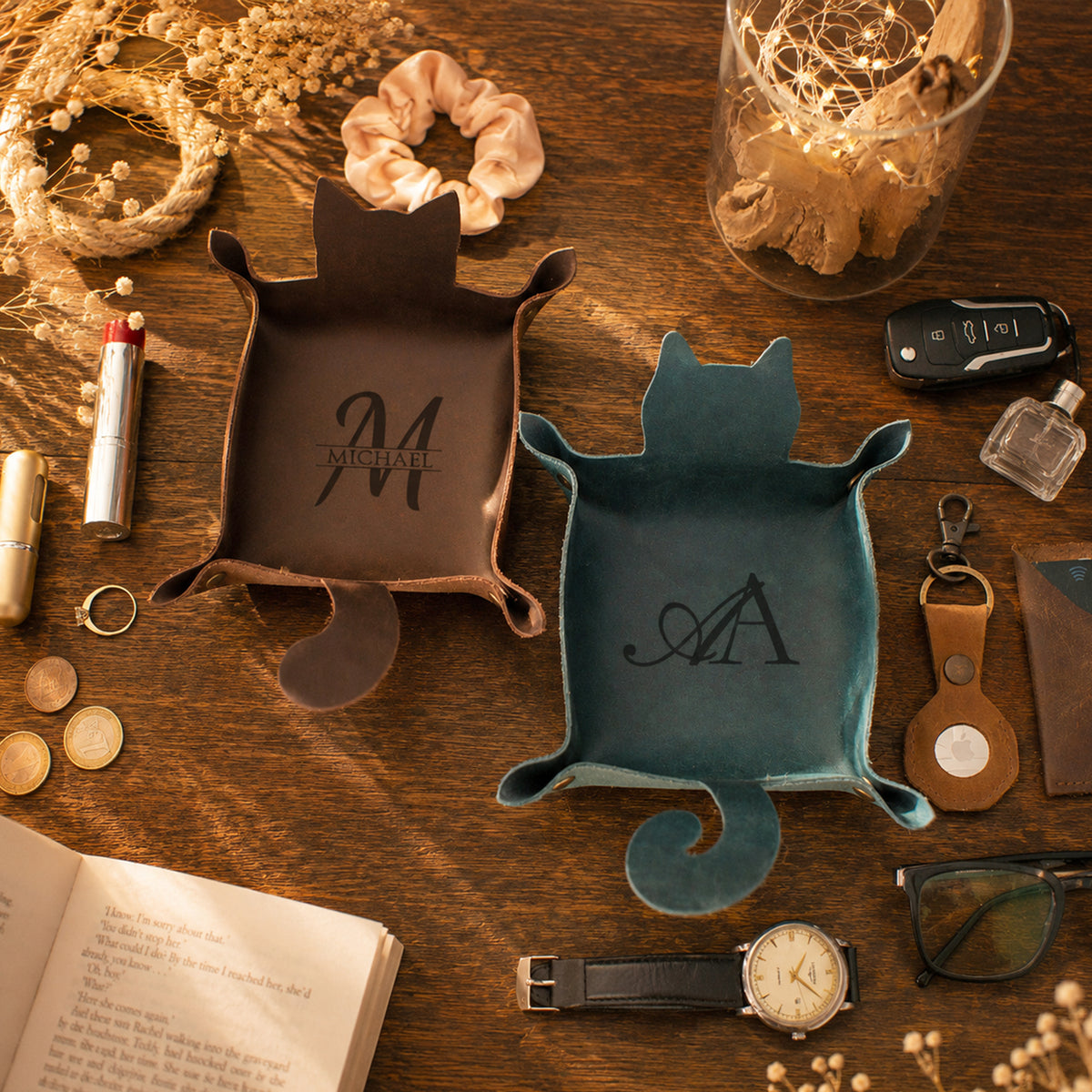 Two ceramic turtle-shaped coasters or trays on a wooden table, one dark brown with the name 'MICHAEL' and one teal blue with the initials 'M & A', surrounded by lifestyle props like a watch, sunglasses, keys, and an open book.