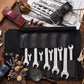 A set of eight silver open-end wrenches with engraved text, displayed in a black leather tool roll on a rustic wooden surface, surrounded by styled props including a magnifying glass, dried flowers, and model cars.