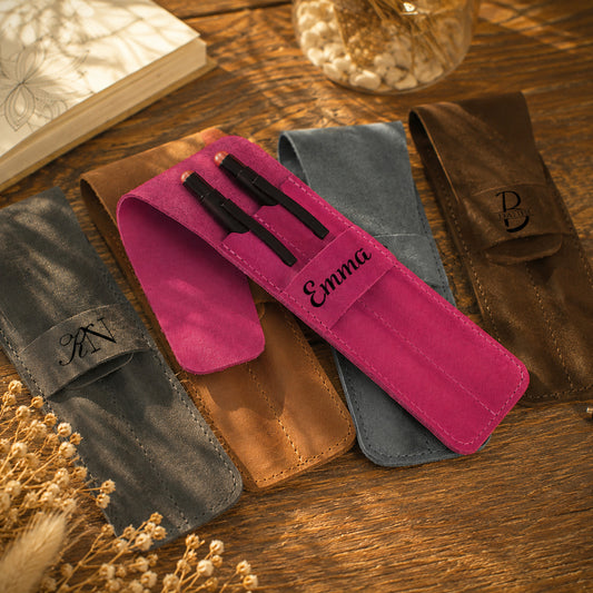 Four personalized pen pouches in different colors (gray, brown, blue, and magenta) arranged on a wooden table with dried flowers and a book in the background. The magenta pouch is open, showing two black pens, and is personalized with the name 'Emma'.