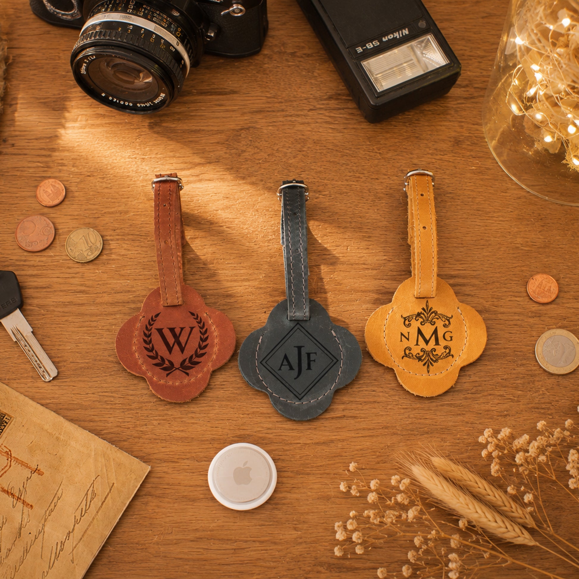 Three personalized leather luggage tags with clover-shaped cutouts and engraved initials are arranged on a wooden tabletop. The tags come in brown, dark gray, and tan colors, styled with vintage props like a camera, coins, a key, and dried flowers.