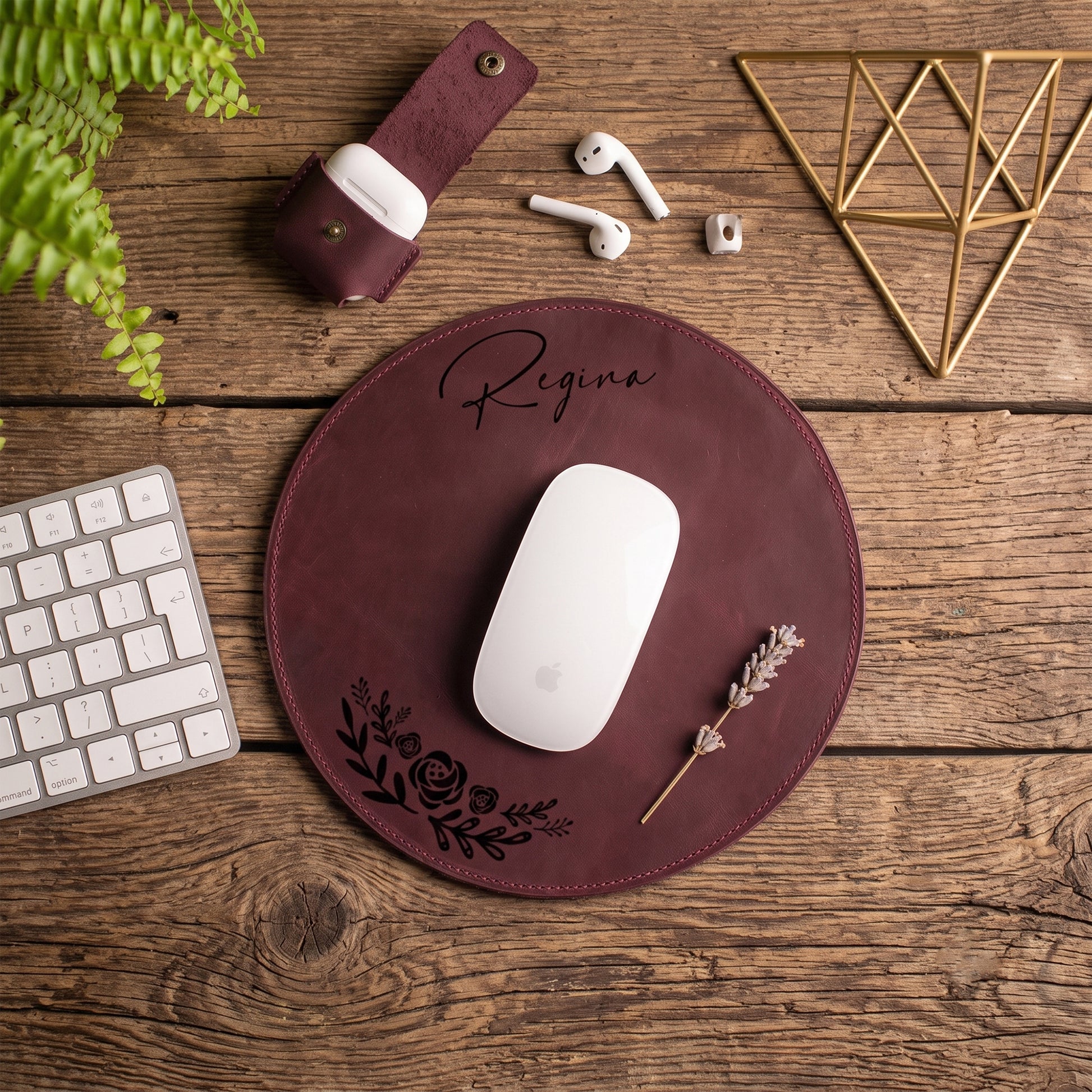 A personalized round burgundy leather mouse pad with 'Regina' and floral engraving, styled on a wooden desk with a white mouse, keyboard, earbuds, and decorative props.