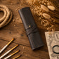 A dark brown leather roll-up case with a snap strap closure, styled on a rustic wooden surface with dried flowers, paintbrushes, and a vintage notebook.