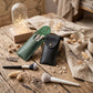 Two personalized makeup pouches, one dark green and one black leather, both engraved with the name 'Kelly', displayed on a rustic wooden table alongside makeup brushes, dried flowers, and a glass dome light.