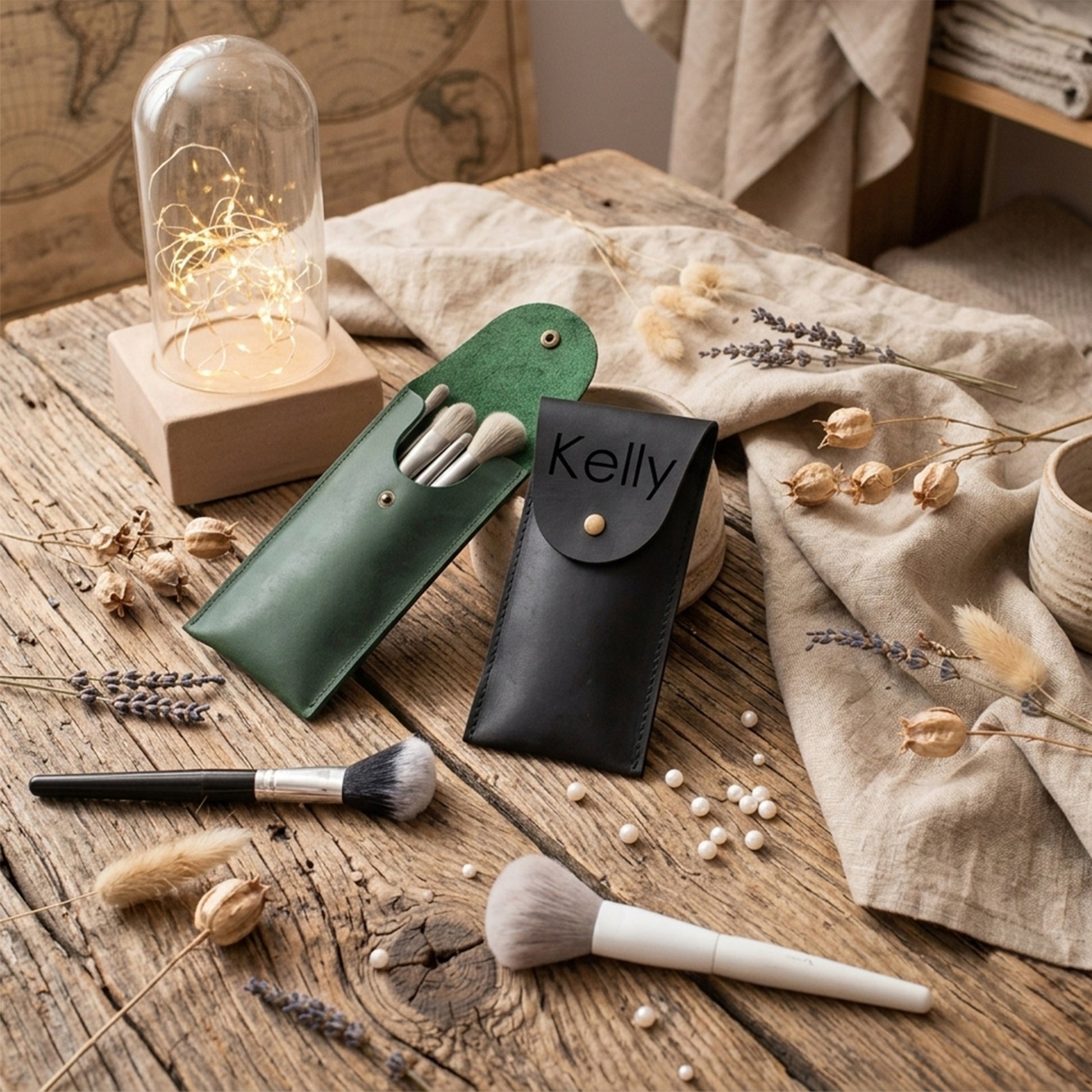 Two personalized makeup pouches, one dark green and one black leather, both engraved with the name 'Kelly', displayed on a rustic wooden table alongside makeup brushes, dried flowers, and a glass dome light.