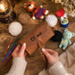 A person holds a personalized brown leather crochet needle case with the name 'De mie' embroidered on the front, surrounded by knitting supplies and handmade toys on a wooden table.