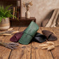 A leather roll-up case in sage green with the words 'Just For Me' printed on it, displayed on a rustic wooden table alongside other leather cases in burgundy, black, and brown, decorated with dried wheat and lavender.