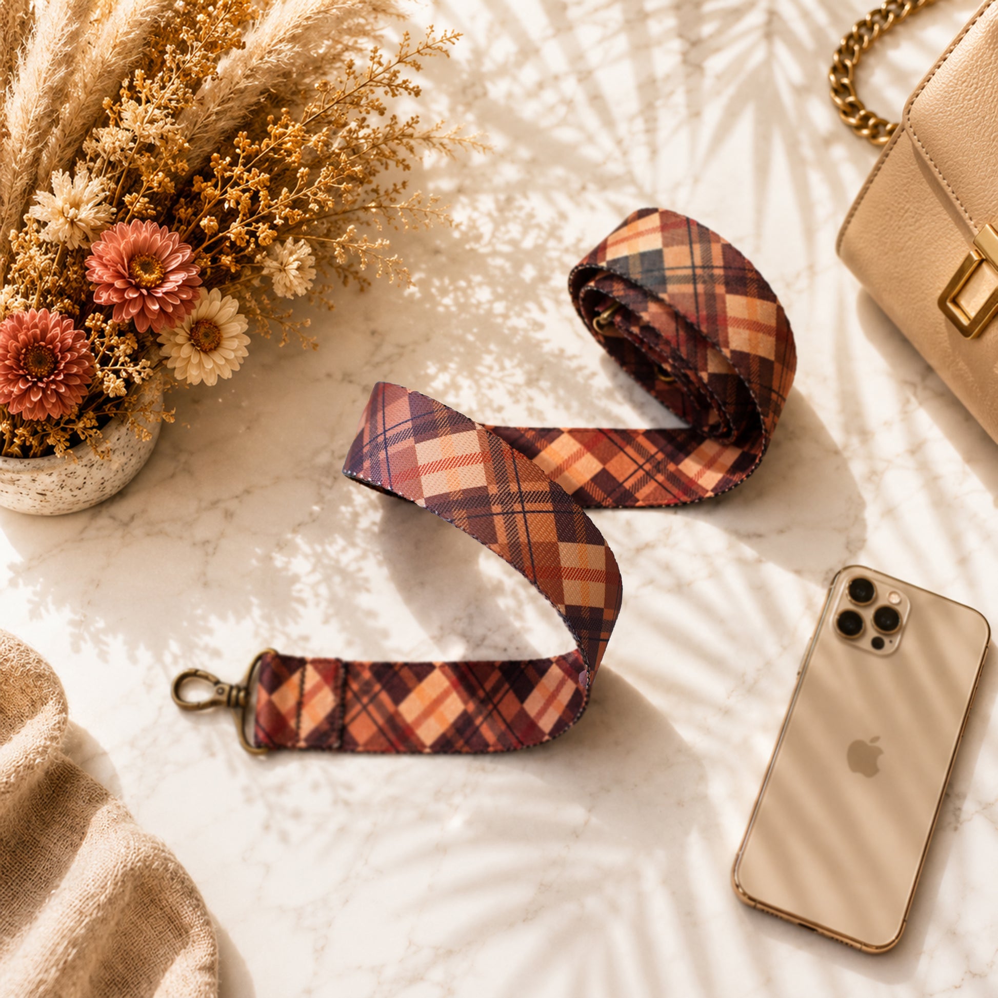 Plaid camera strap on a marble surface with a phone, flowers, and handbag.