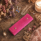 A bright pink velvet jewelry wallet with two gold snap buttons, styled on a rustic wooden table surrounded by dried flowers, jewelry, rings, and a lit candle.
