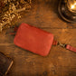 A reddish-brown leather crossbody pouch with a strap, laid flat on a rustic wooden table with dried flowers and a vintage lamp in the background.