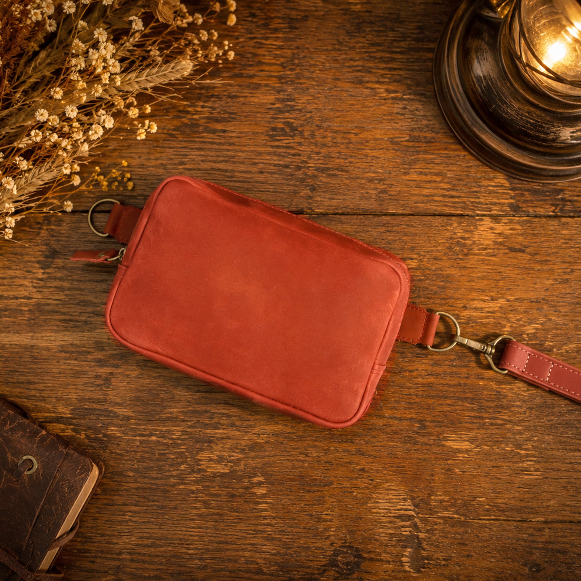 A reddish-brown leather crossbody pouch with a strap, laid flat on a rustic wooden table with dried flowers and a vintage lamp in the background.