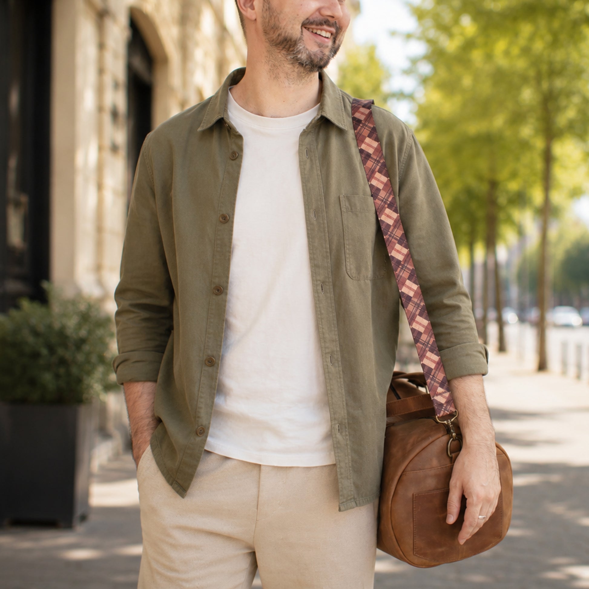 Man wearing a green shirt and beige pants, holding a brown leather bag with a patterned strap on a street.