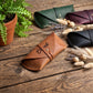 A tan leather glasses case with an engraved script logo is displayed on a rustic wooden table, surrounded by other color options (green, burgundy, black) and decorative dried grasses and a potted fern.