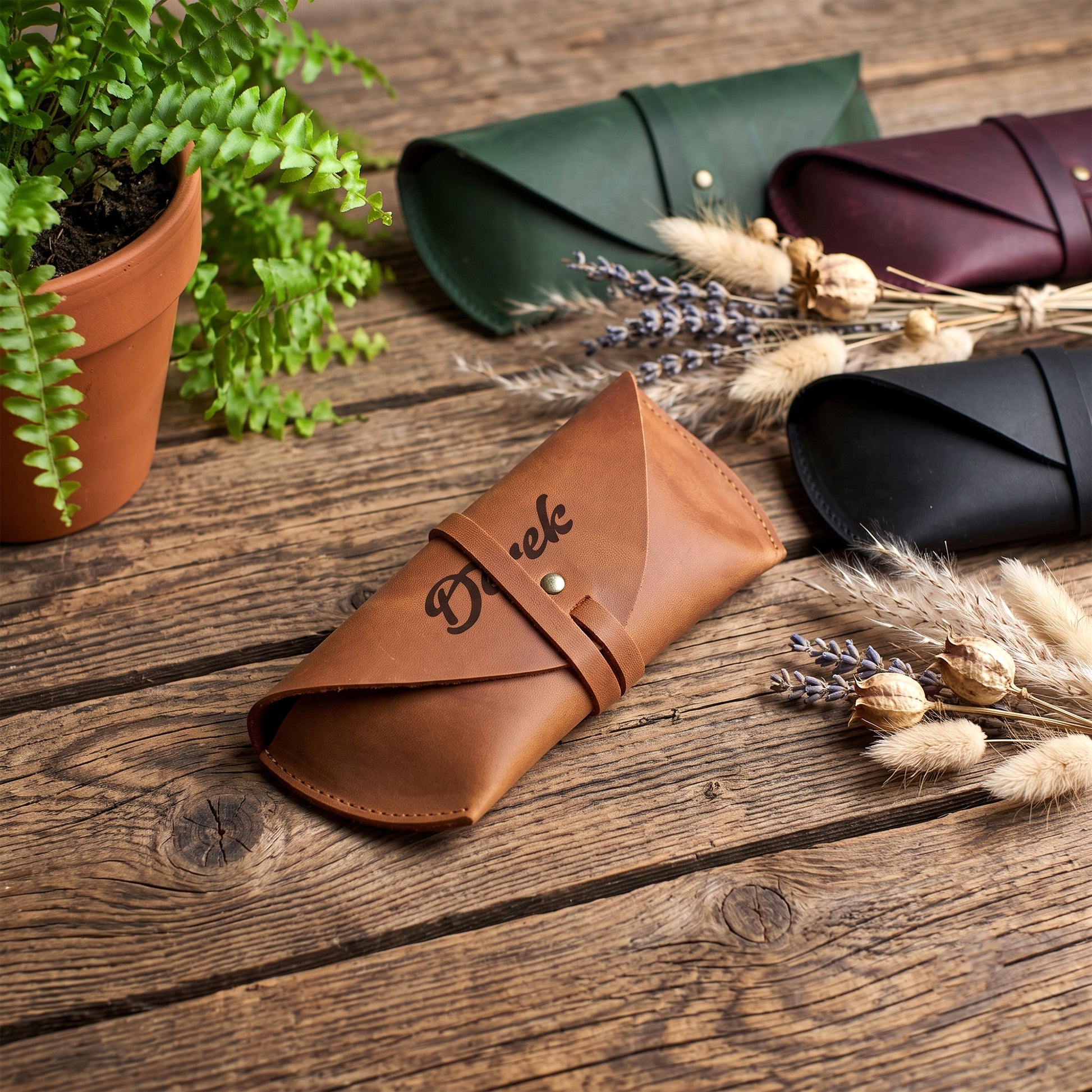 A tan leather glasses case with an engraved script logo is displayed on a rustic wooden table, surrounded by other color options (green, burgundy, black) and decorative dried grasses and a potted fern.