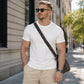 Man wearing a white t-shirt and beige pants with a crossbody bag on a city street.