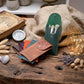 A set of leather makeup brush pouches in green and brown, shown with makeup brushes on a rustic wooden table surrounded by dried flowers, a compass, and warm fairy lights.