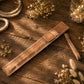 A tan leather sheath or sleeve with a strap, laid diagonally on a rustic wooden surface surrounded by dried flowers and warm string lights.