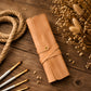 A rolled tan leather or faux-leather travel case with a snap strap closure, styled on a rustic wooden surface with dried flowers, rope, and paintbrushes.