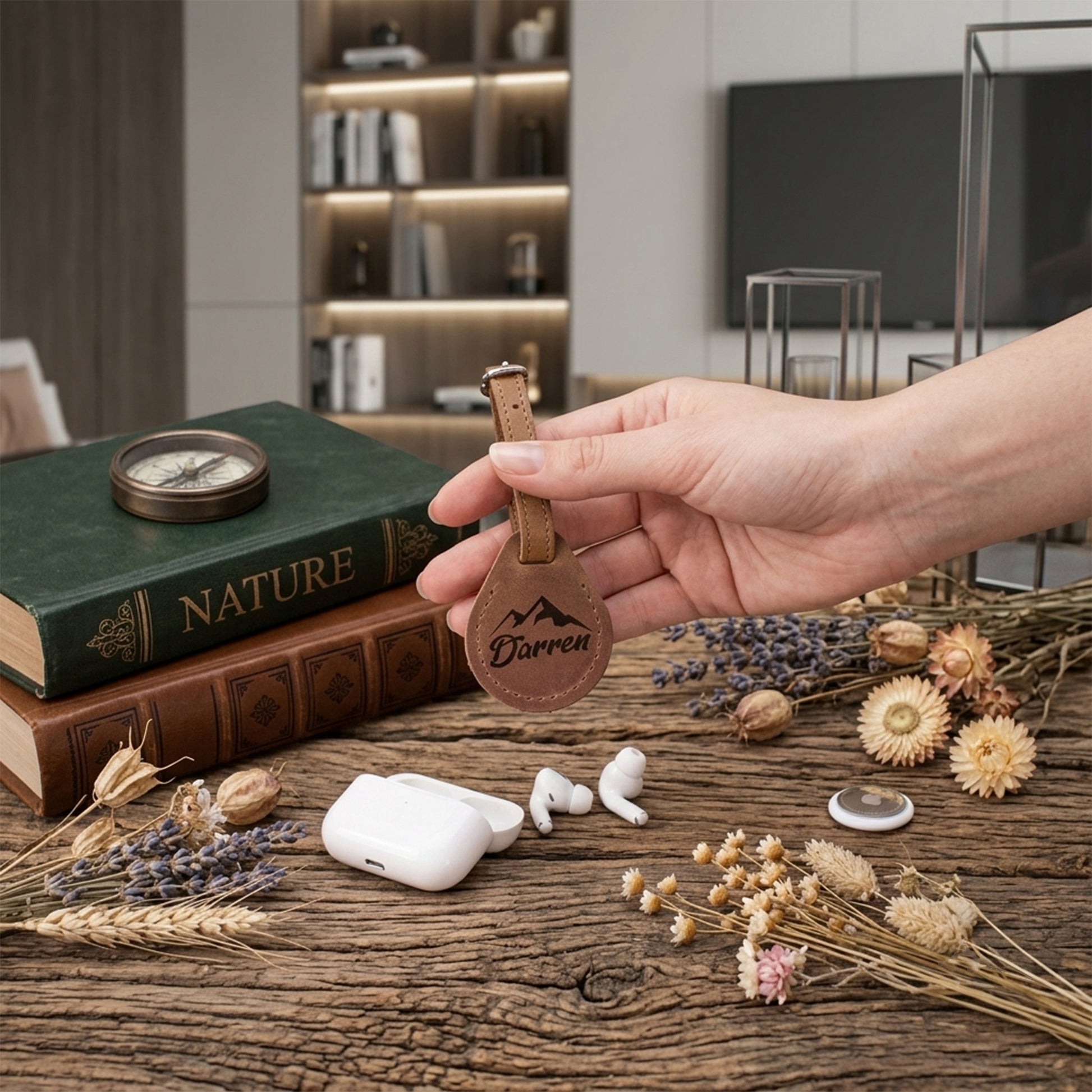 A hand holds a brown leather keychain with the name 'Darren' engraved on a round tag, shown in a styled indoor setting with books, dried flowers, and white wireless earbuds nearby.