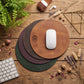 Three round leather mouse pads in tan, dark brown, and forest green are stacked on a rustic wooden desk, with a white computer mouse on the top tan pad. The scene is styled with a keyboard, earbud case, white earbuds, a leather wallet, dried flowers, and a plant.