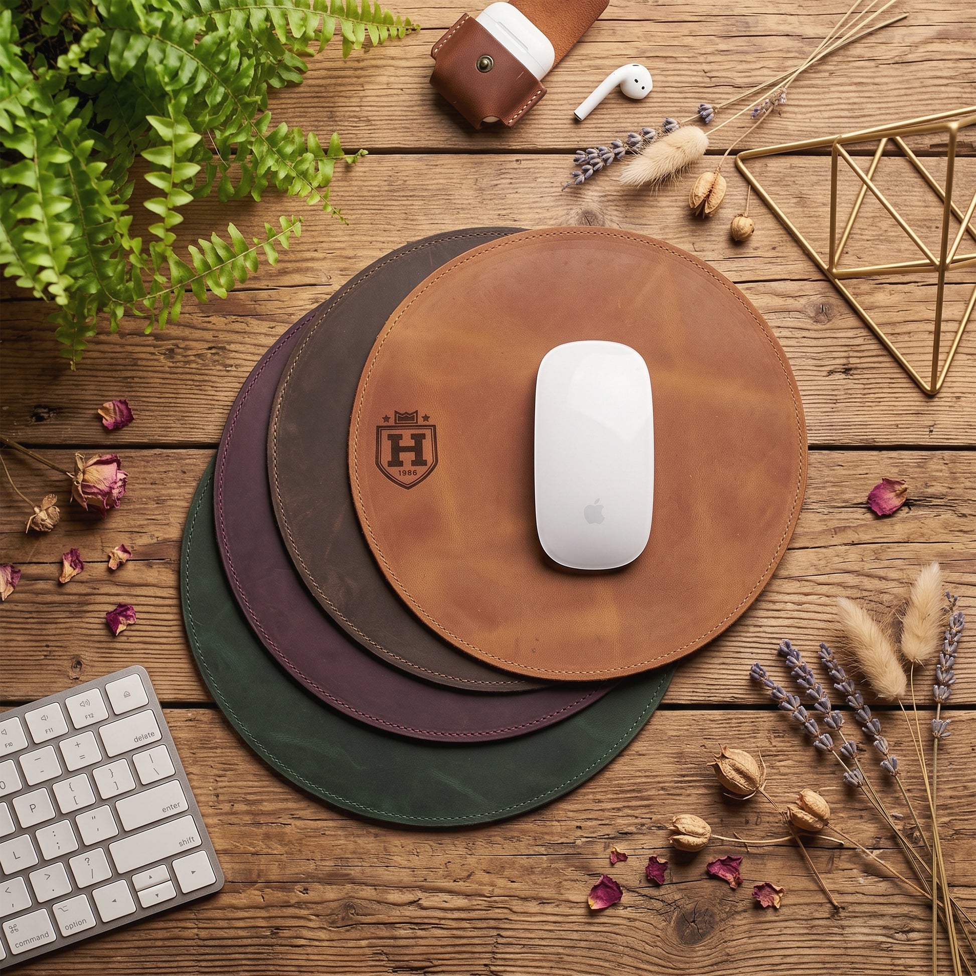 Three round leather mouse pads in tan, dark brown, and forest green are stacked on a rustic wooden desk, with a white computer mouse on the top tan pad. The scene is styled with a keyboard, earbud case, white earbuds, a leather wallet, dried flowers, and a plant.
