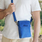 Person holding a blue crossbody bag with 'David' embroidered on it, outdoors.
