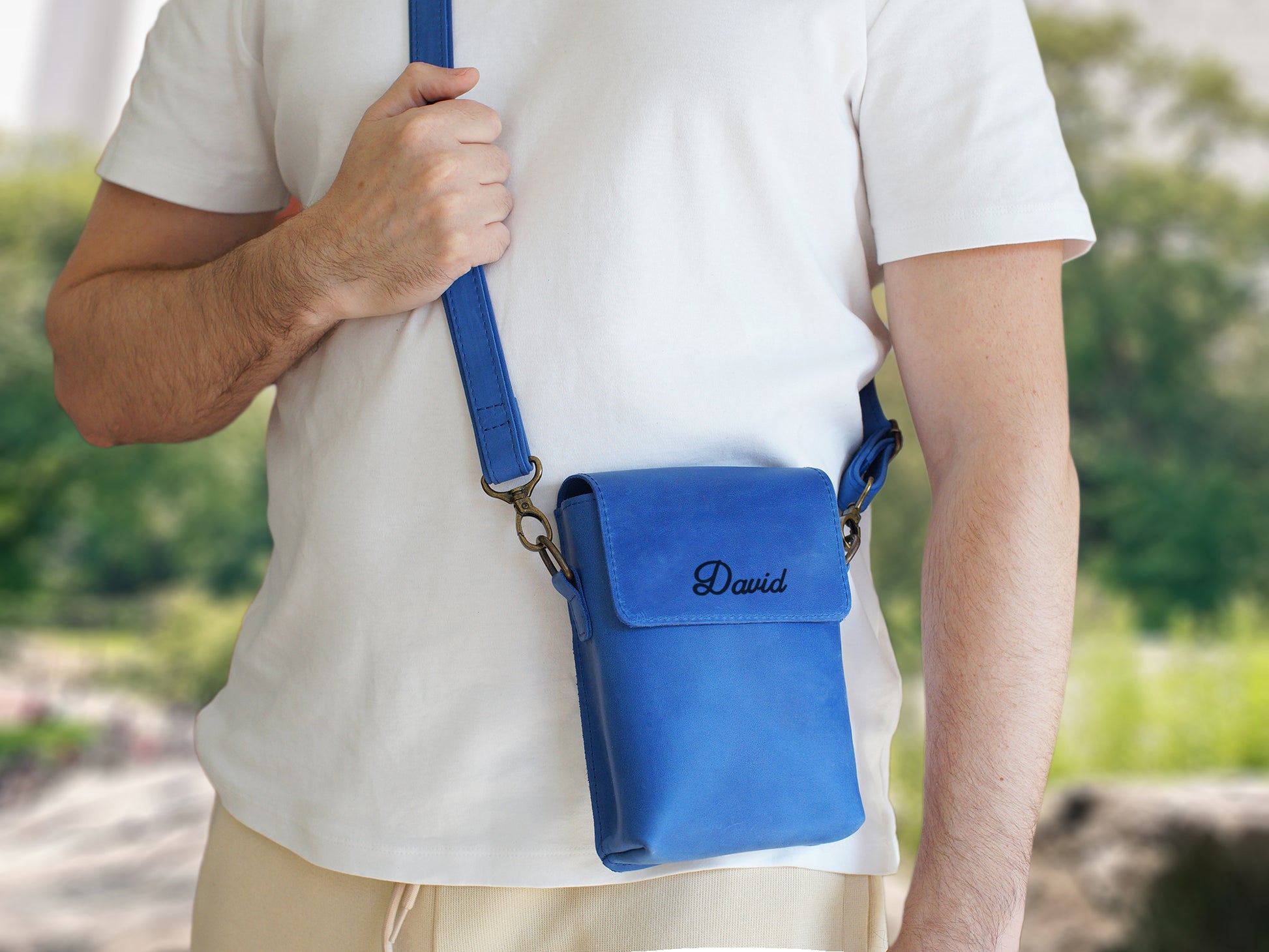 Person holding a blue crossbody bag with 'David' embroidered on it, outdoors.
