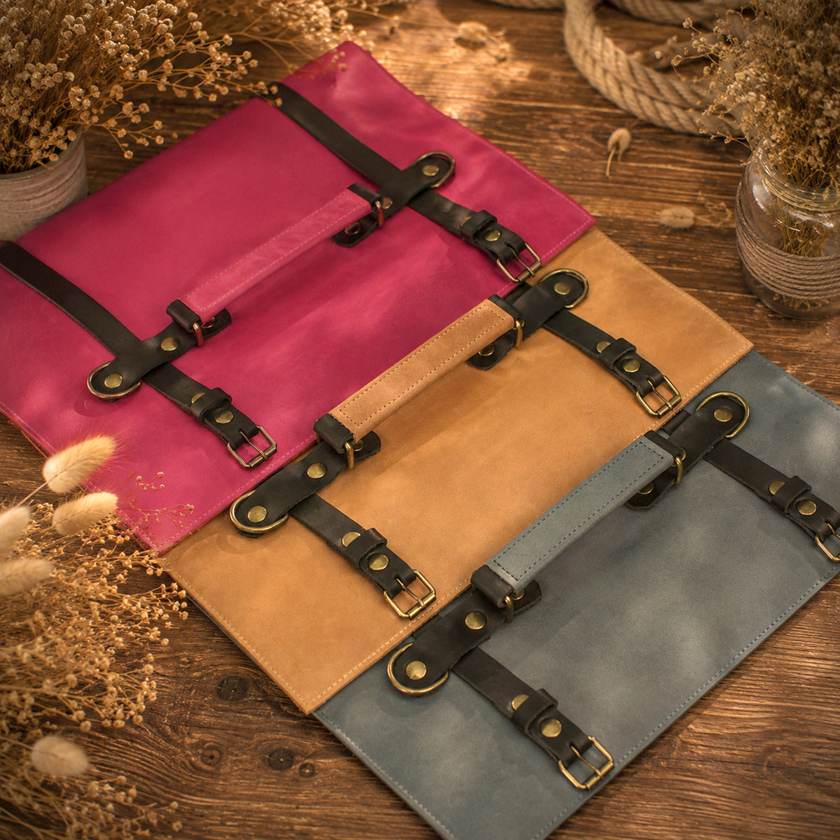 Three leather document pouches with black buckle straps are arranged diagonally on a rustic wooden surface with dried flowers and rope props. The pouches are in magenta pink, tan, and slate blue colors.