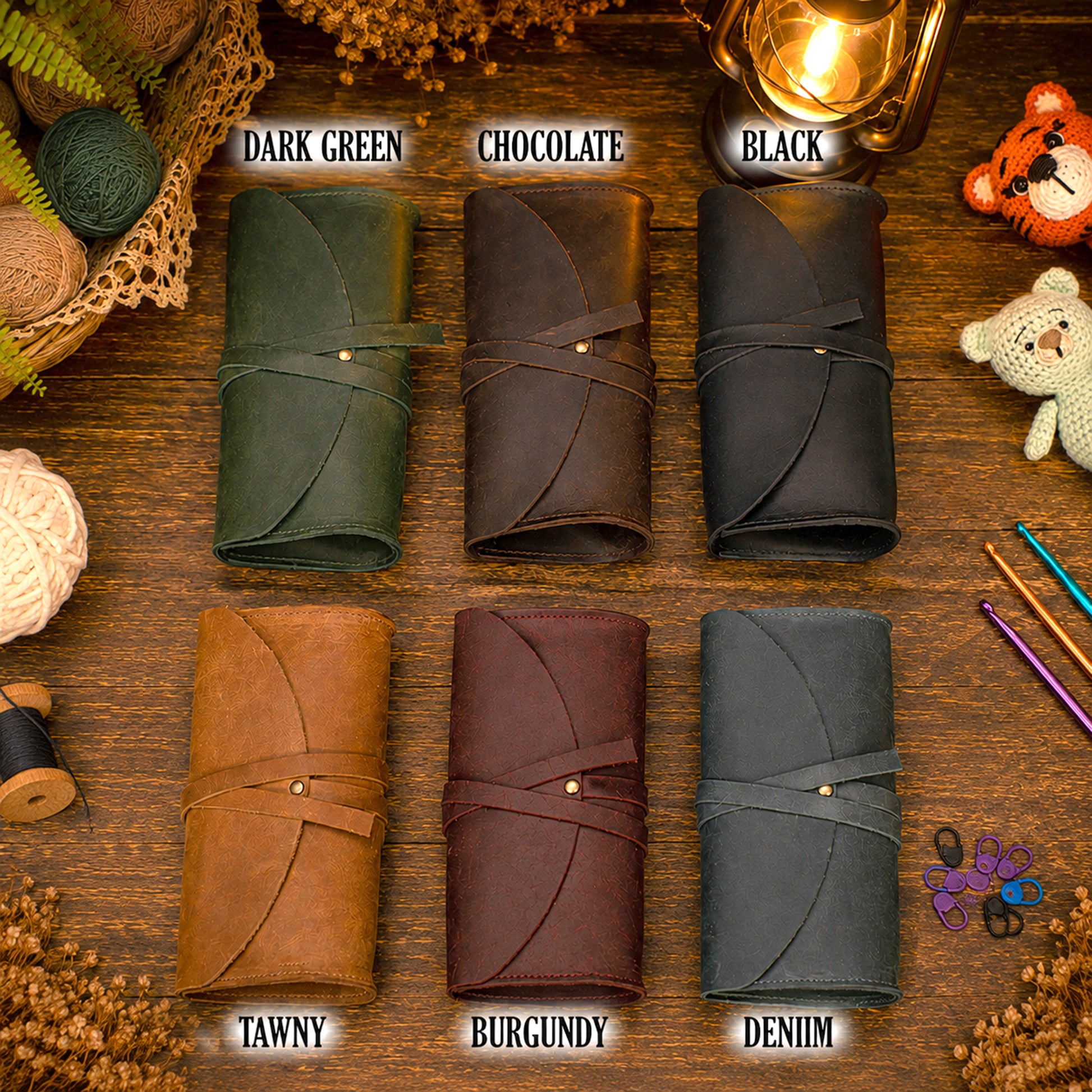 Six rolled leather journal covers arranged in a 2x3 grid on a wooden surface, labeled Dark Green, Chocolate, Black, Tawny, Burgundy, and Denim. Decorative craft props and a warm lantern appear around the products.