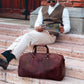 Man sitting on steps with a brown leather duffel bag next to him, holding a newspaper.
