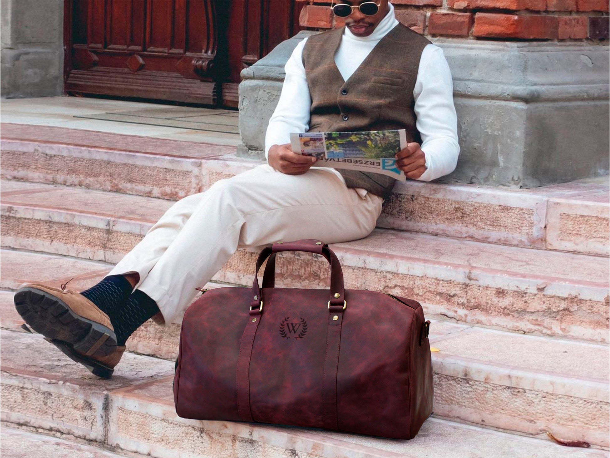 Man sitting on steps with a brown leather duffel bag next to him, holding a newspaper.