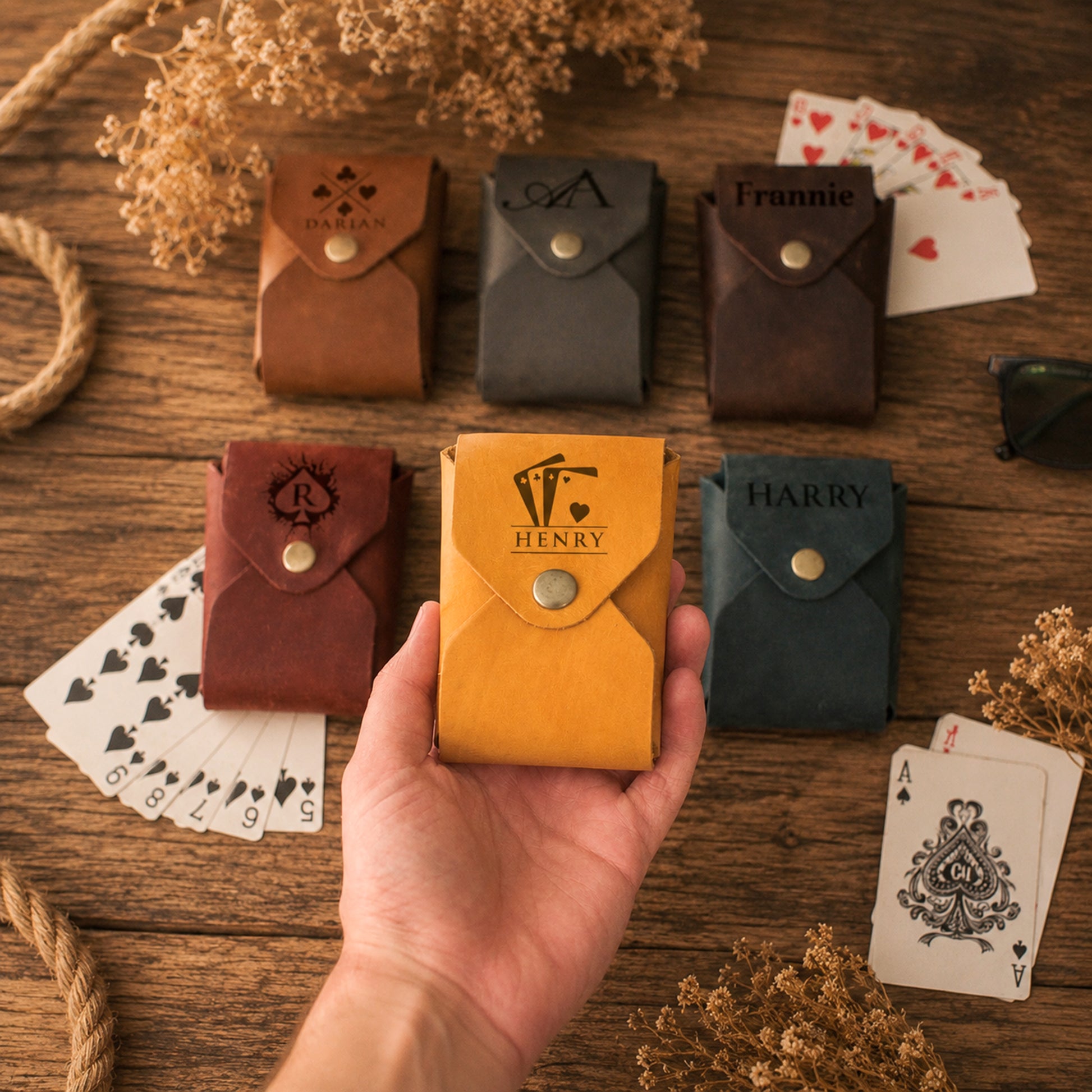 A hand holds a yellow personalized leather card holder with the name 'HARRY' and a monogram, surrounded by five other matching leather card holders in brown, gray, and dark blue on a wooden tabletop with playing cards and dried flowers.