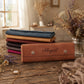A stack of folded leather accessories, including a prominent brown envelope-style case engraved with the name 'Abigail', is displayed on a rustic wooden table alongside dried flowers and jewelry in a softly lit indoor setting.