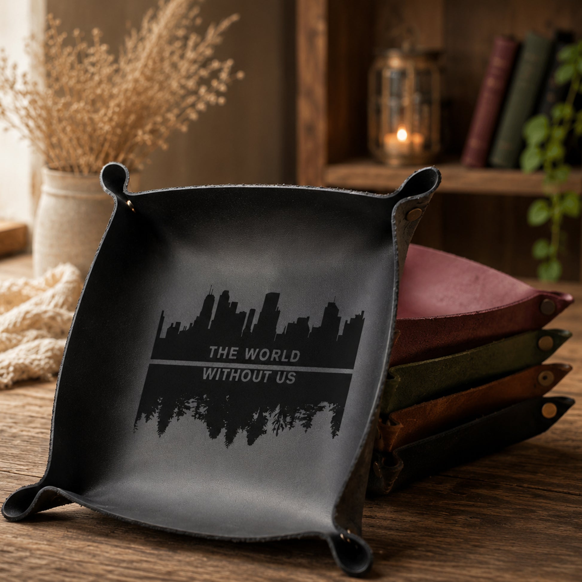 A black leather square tray with rounded corners and a printed black skyline graphic and the text 'THE WORLD WITHOUT US' sits on a wooden table. A stack of similar leather trays in muted pink, green, brown, and black is visible behind it, with a softly blurred rustic interior background including books, a candle, and dried flowers.