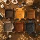 Six personalized leather tray coasters arranged in a flat-lay on a wooden table with various lifestyle props. The coasters feature different colors (dark brown, mustard yellow, medium brown, silver, copper, and navy) and are customized with monogram names or initials.