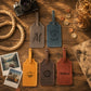 Four personalized leather luggage tags arranged on a rustic wooden table with travel-themed props including a camera, rope, coins, instant photos, and a map. The tags are shown in gray, blue, yellow, brown, and reddish-brown colors, each engraved with a monogram or name.