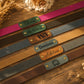 Six personalized leather straps in different colors arranged diagonally on a rustic wooden surface. Each strap has a rectangular metal nameplate engraved with a personalized name or word, such as 'Eliana', 'Adam', 'Eva', 'Dominic', 'Adeline', and 'yoga'.