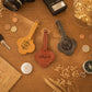 Three personalized leather luggage tags with cloud-shaped cutouts are arranged on a wooden tabletop with styled props including a camera, coins, a notebook, dried flowers, and a glass of warm lights. The tags are in mustard yellow, reddish-brown, and dark gray, each engraved with a monogram or name.