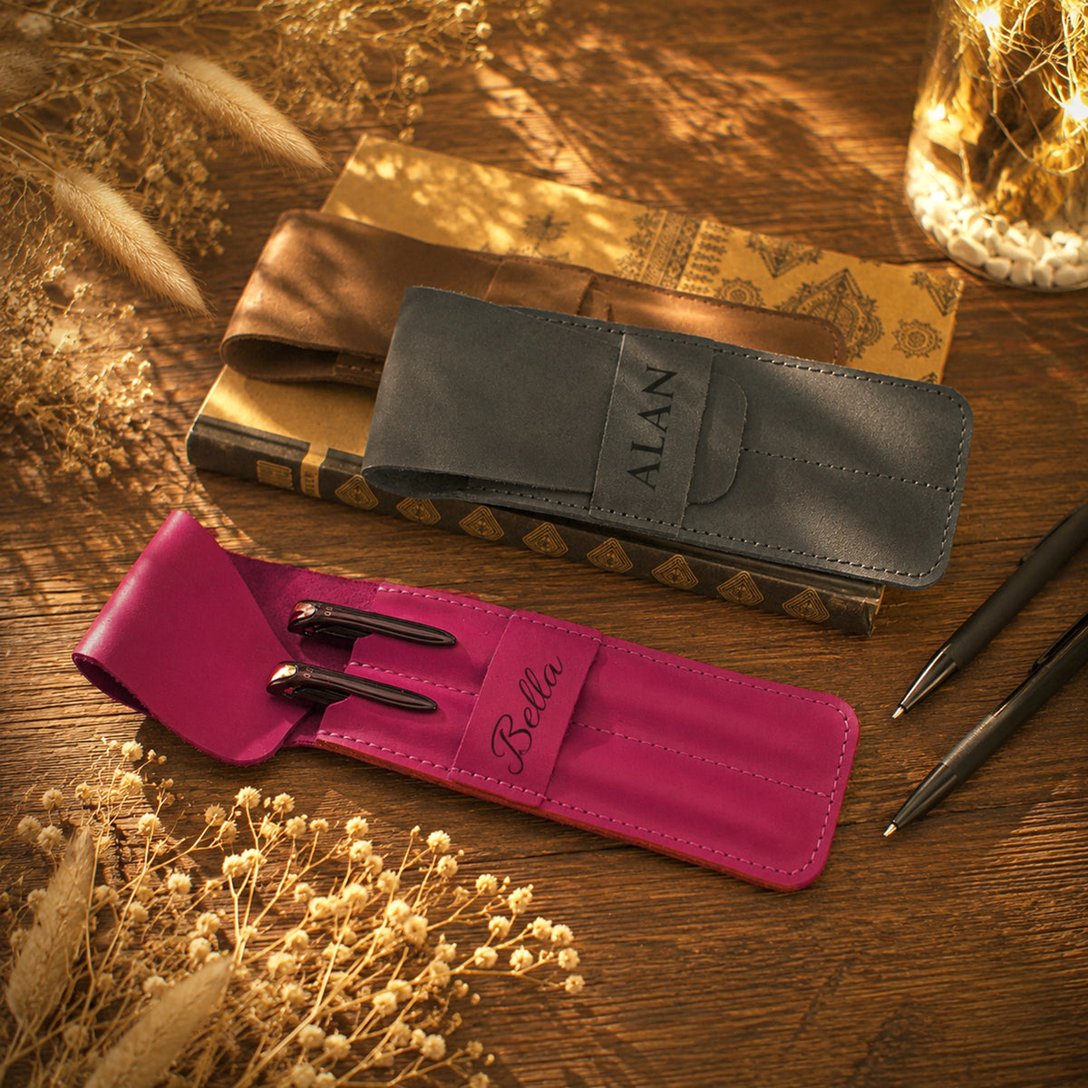 Two personalized leather pen cases on a wooden table, one in dark gray with the name 'ALAN' and one in bright pink with the name 'Bella', both styled with pens and dried grasses.