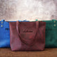 Three tote bags in blue, red, and green with embroidered names on a wooden surface.