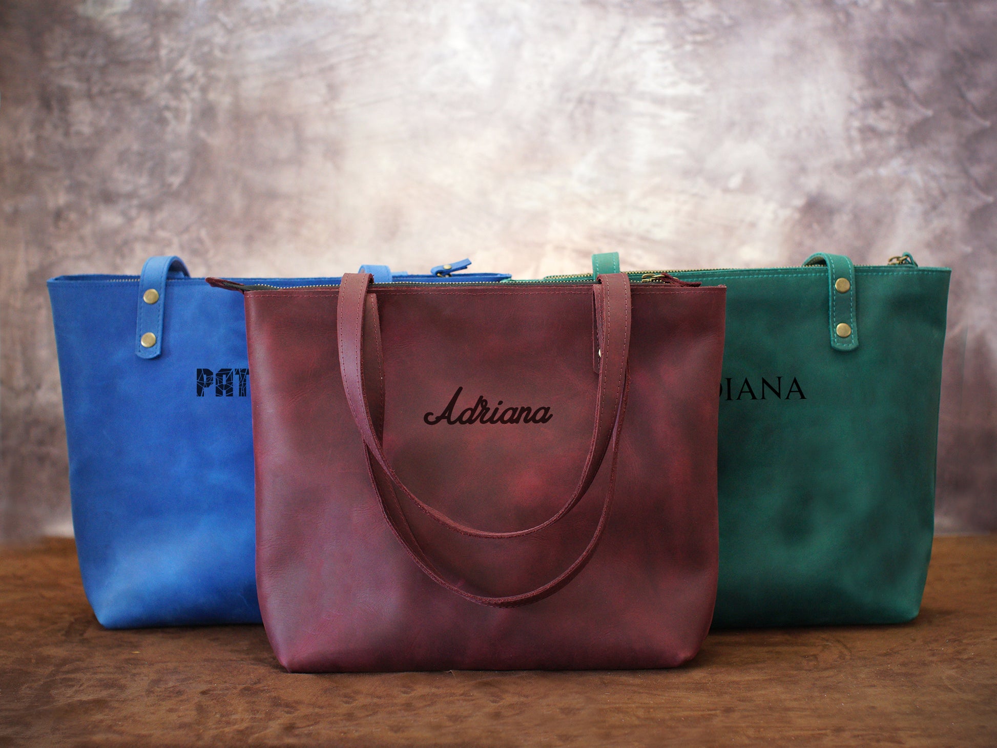 Three tote bags in blue, red, and green with embroidered names on a wooden surface.